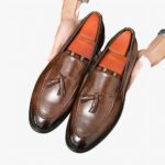 Des mocassins marrons en cuir à bout pointus et foncés avec des glands décoratifs dans les mains d'un homme qui les tient au-dessus d'un parquet en bois.