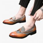 Des mocassins bi-matière avec un motif à chevron blanc et noir et du cuir marron clair et des glands décoratifs aux pieds d'un homme portant un pantalon de costume noir.