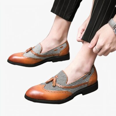 Des mocassins bi-matière avec un motif à chevron blanc et noir et du cuir marron clair et des glands décoratifs aux pieds d'un homme portant un pantalon de costume noir.