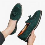 Des mocassins en daim vert émeraude avec glands décoratifs au pied à dans la main d'une personne qui se tient assise sur un sol gris clair.