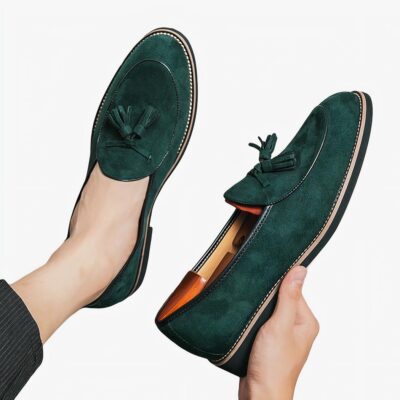 Des mocassins en daim vert émeraude avec glands décoratifs au pied à dans la main d'une personne qui se tient assise sur un sol gris clair.