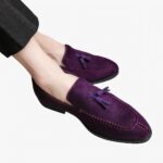 Des mocassins violets en daim avec glands décoratifs, aux pieds d'un homme aux jambes croisées portant un pantalon de costume noir à rayures fines blanches.