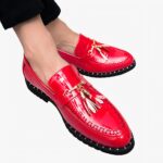 Des mocassins vernis rouge vif avec des glands décoratifs sur les pieds d'un homme se tenant assis les jambes croisées et portant un pantalon de costume noir.