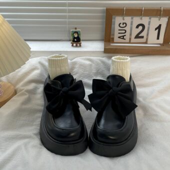 Une paire de mocassins en cuir lisse noir posée sur un lit blanc, un grand noeud décore le dessus et des petites chaussettes beiges remplissent la chaussure.