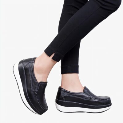 Photo de mocassins orthopédiques à plateforme noirs avec une semelle blanche, portée par des jambes de femme en pantalon noir.