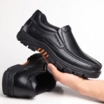 Photo d'une paire de mocassins orthopédiques en cuir noir à semelle épaisse, une chaussure est couchée sur le sol, l'autre est tenue dans une main.