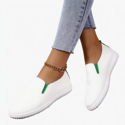 Sur un sol parquet gris, une paire de mocassins blanc perforé avec petits élastiques verts sur les cotés. Ils sont portés pieds nus avec un jean bleu clair et une chaine à la cheville.