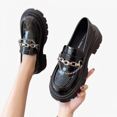 deux mocassins noirs en simili cuir verni avec chaînes décoratives sur le devant, à semelle épaisse, un porté par une jambe, l'autre portée par une main, sur fond d'un mur beige