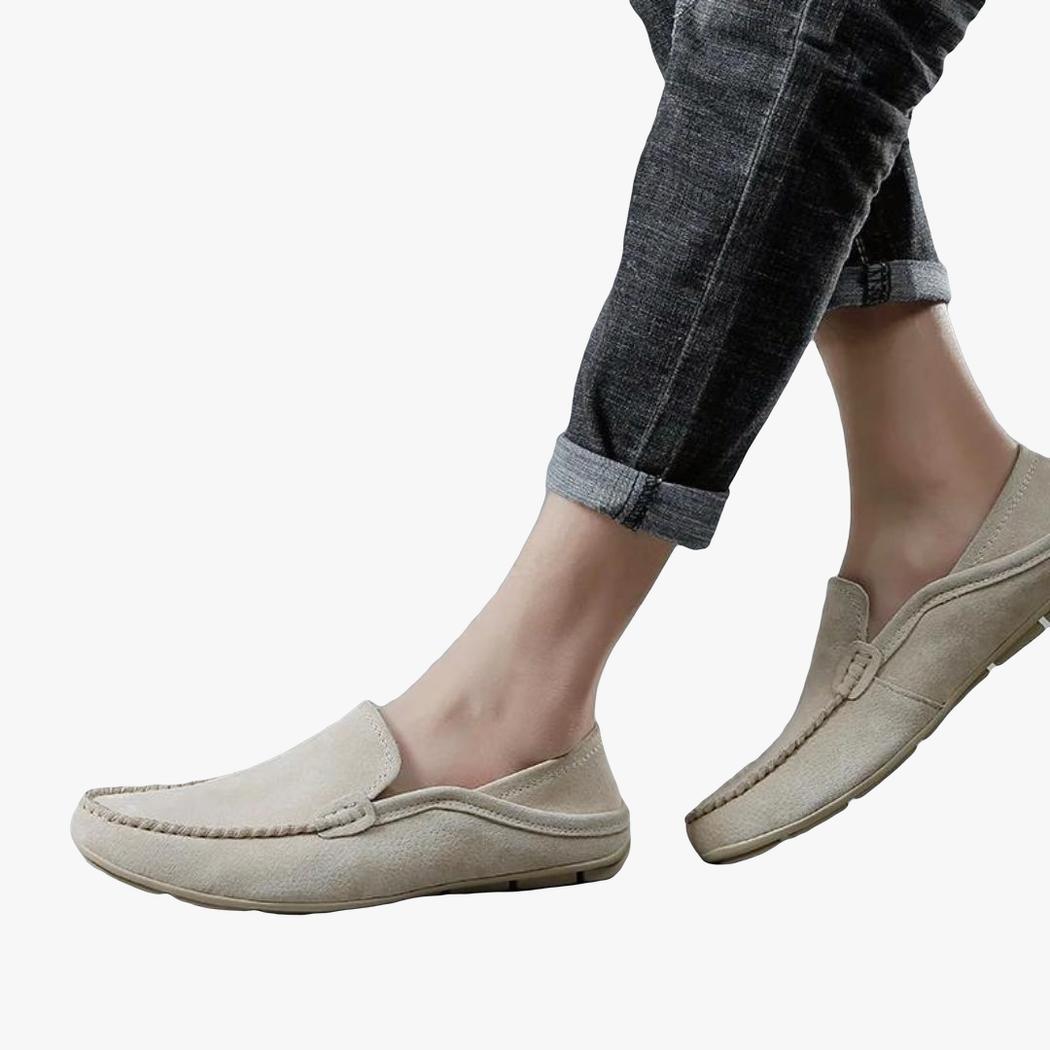 on voit sur un fond gris clair le bas des jambes d'un homme en jean et on voit ses mocassins beige en cuir véritable