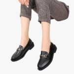 on voit les pieds d'une femme assise qui porte des mocassins noirs en simili cuir avec bouche en métal et talon carré