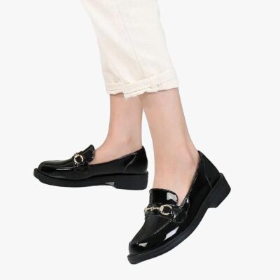 on voit les pieds d'une femme sur un fond blanc qui porte des mocassins vernis en simili cuir noirs avec un talon carré