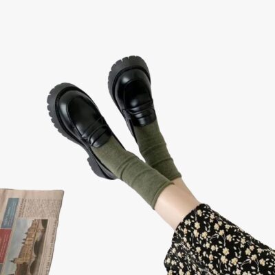 on voit les pieds d'une femme par terre, elle porte des chaussettes kakis et des mocassins noirs à talon carré et semelle épaisse pour femme