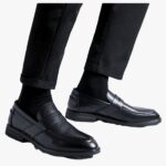 on voit sur un fond clair le bas des jambes d'un homme il porte un pantalon noir et des mocassins noirs classiques en cuir synthétique pour homme