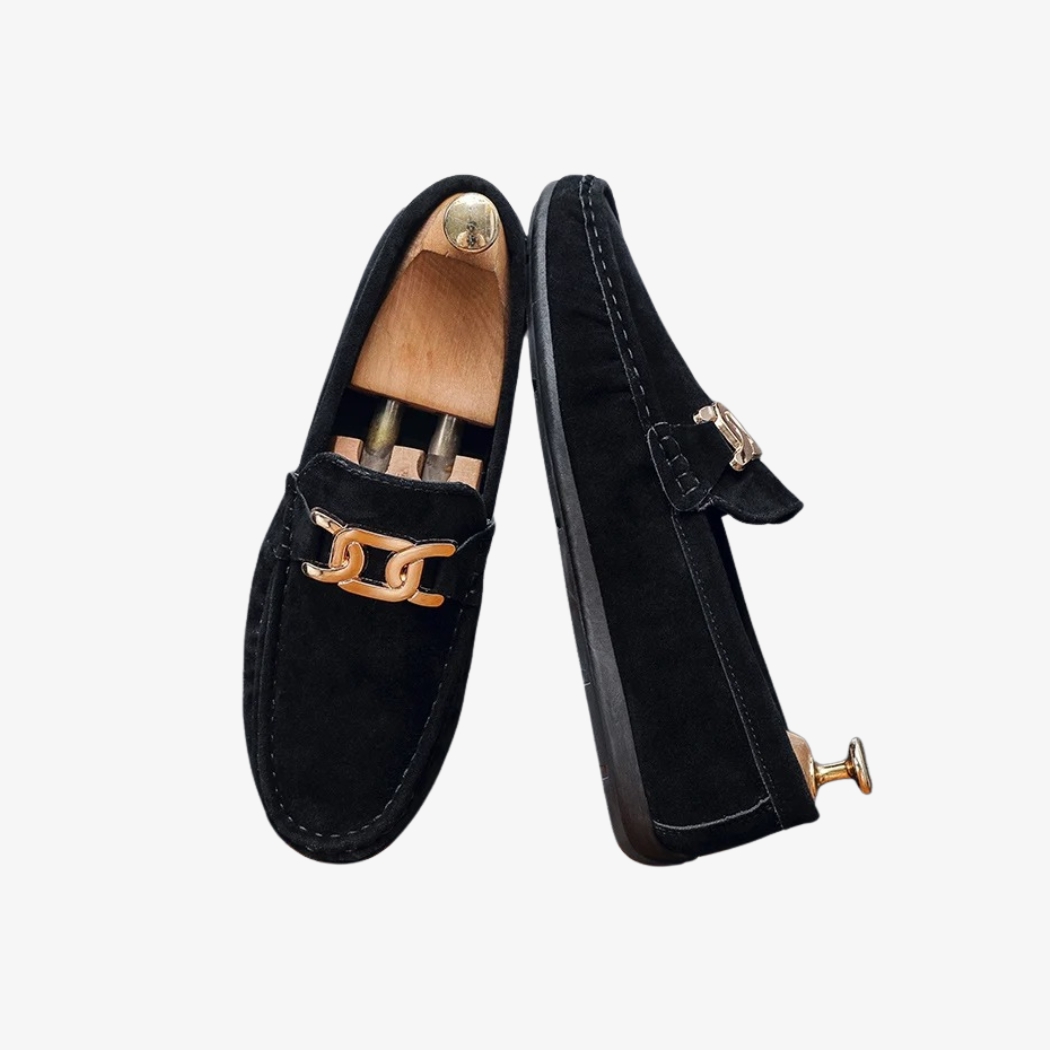 Mocassins en daim noir pour homme, élégants et modernes, ornés d'une boucle métallique dorée sur le dessus. Leur design raffiné met en valeur des coutures visibles qui ajoutent une touche de caractère à l'ensemble. La semelle noire complète leur apparence sobre et chic.